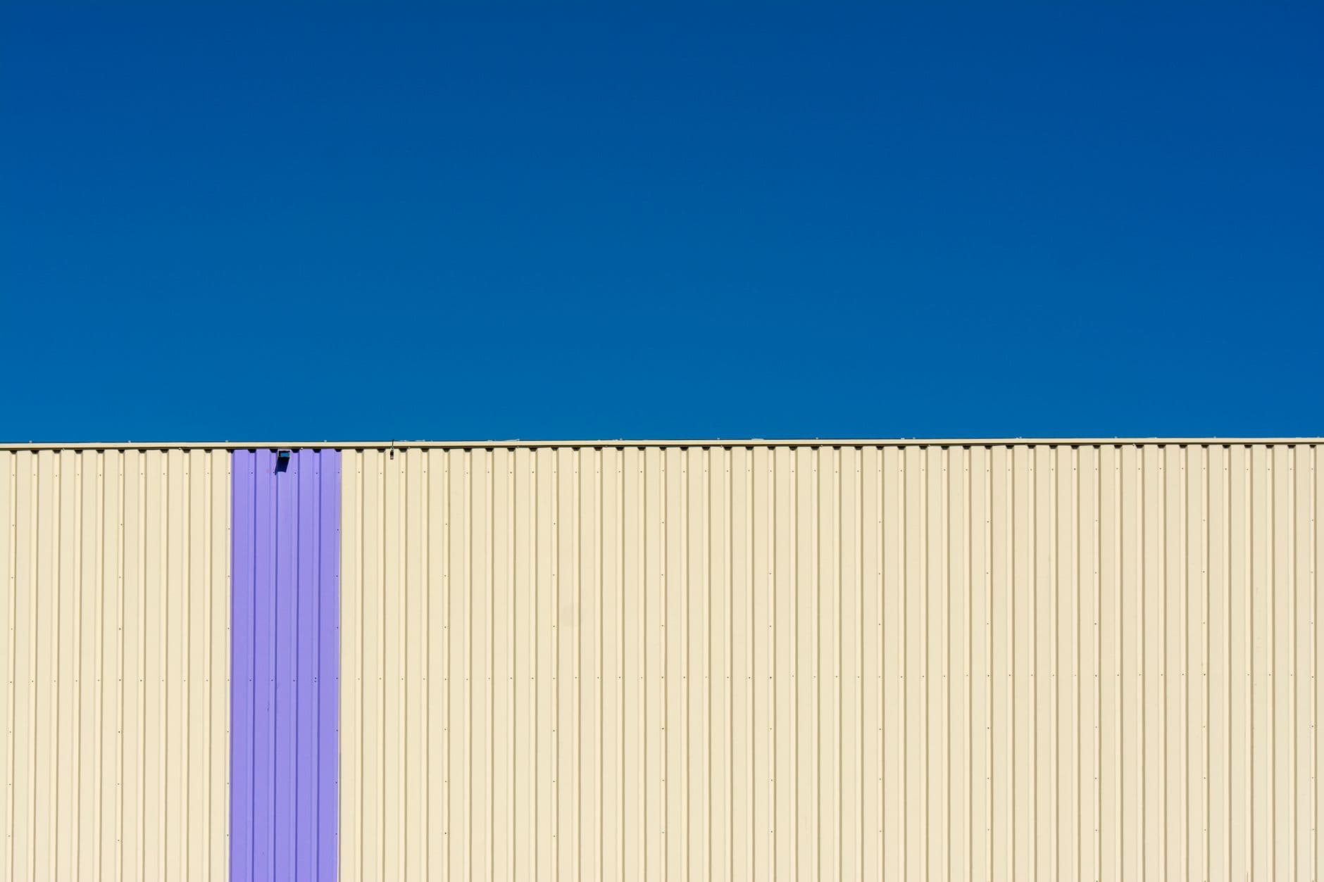 Corrugated cream metal cladding with a single lavender vertical band beneath a clear blue sky.