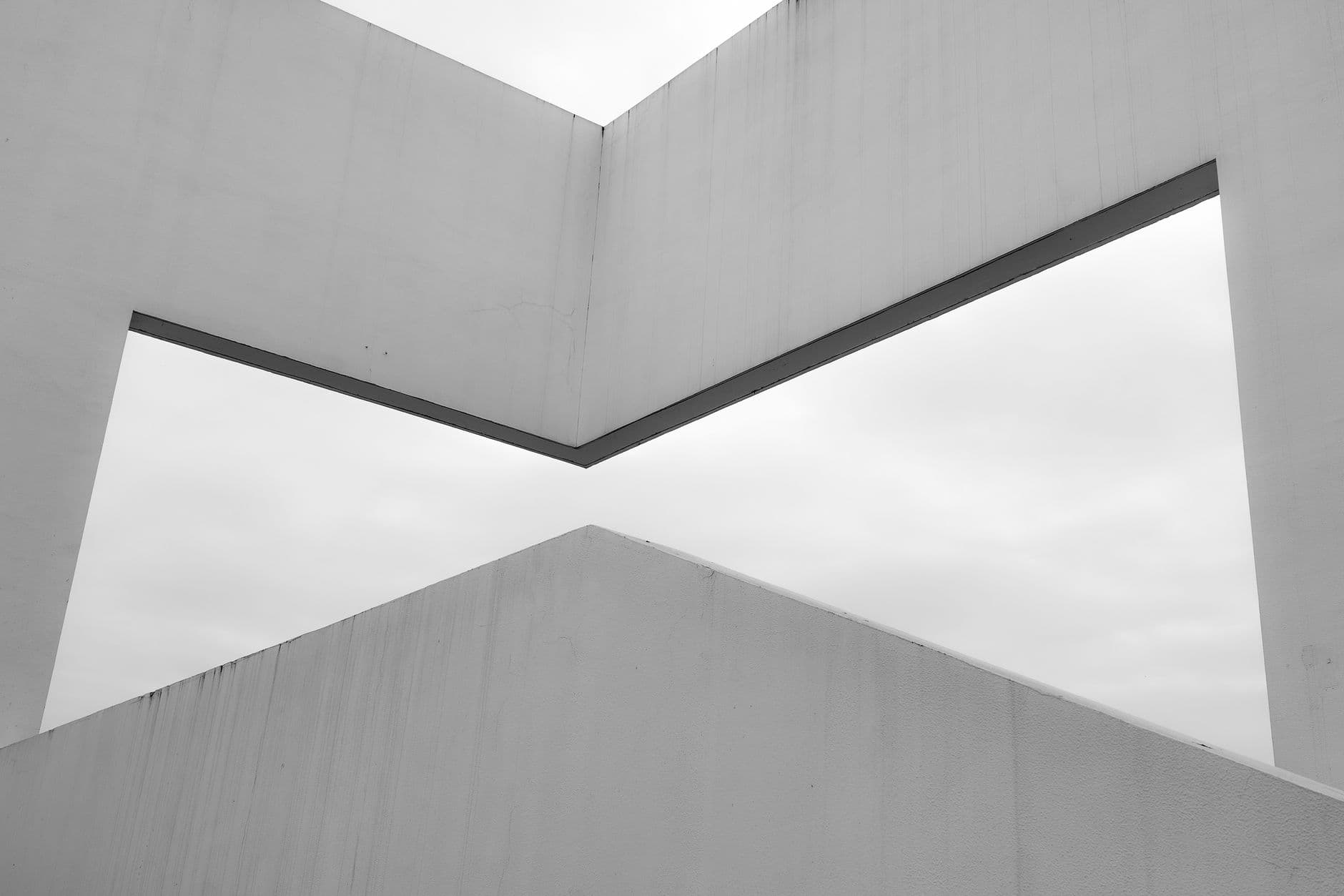 Black-and-white study of intersecting white concrete volumes framing a wedge of sky.