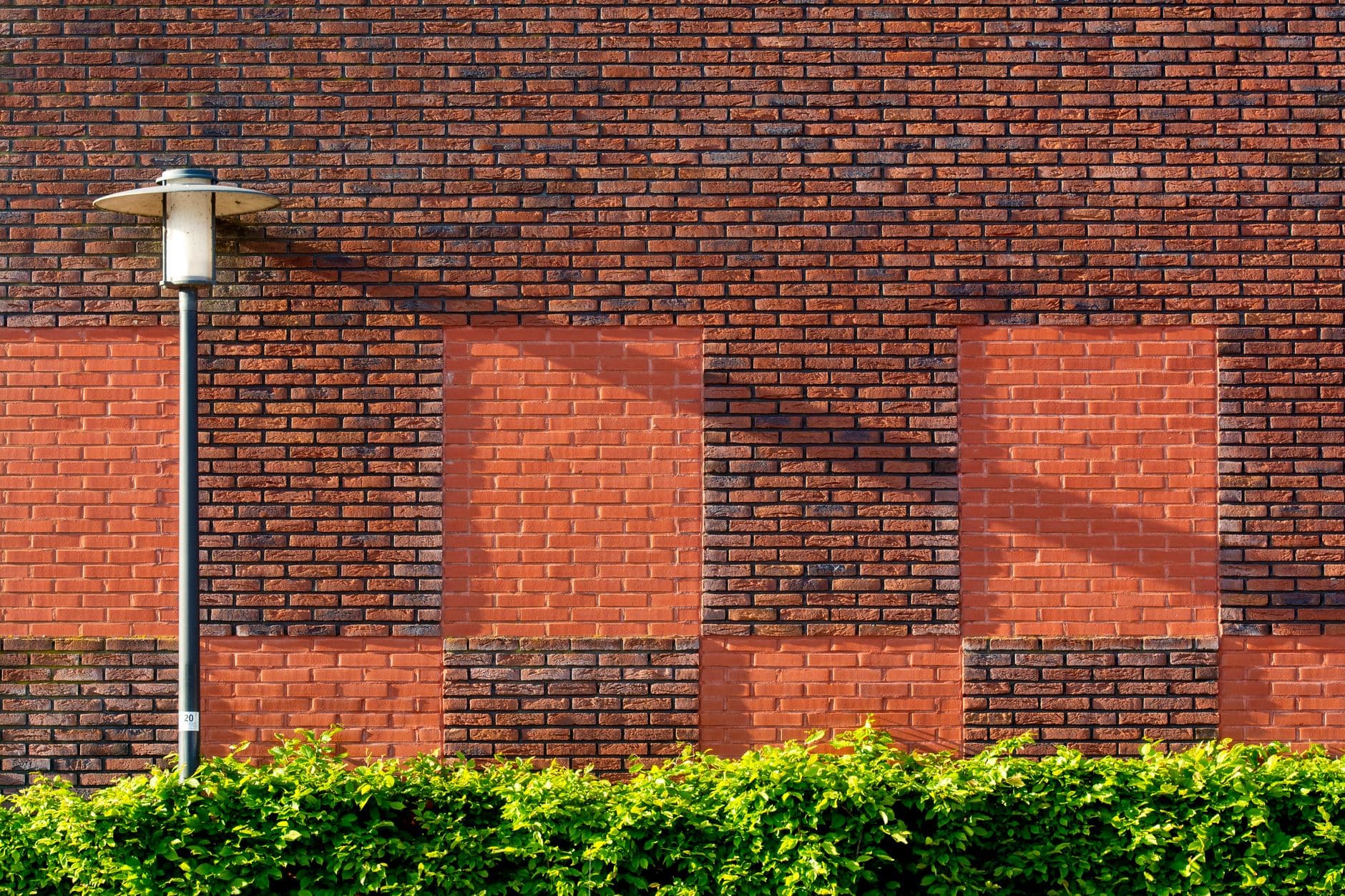 Two-tone red brick wall with recessed blind window panels, a single streetlamp, and a trimmed hedge along its base in low sun.